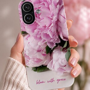 Könnte beinhalten: Eine Handyhülle mit einem Nahaufnahme-Motiv von rosa Pfingstrosen und grünen Blättern. Die Hülle hat eine weiße Basis mit den Worten "bloom with grace" in einer Schreibschrift. Die Handyhülle wird von einer Person gehalten.