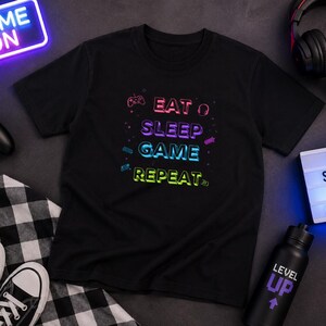 Könnte beinhalten: Ein schwarzes kurzärmeliges T-Shirt mit einem bunten Grafikdruck, der den Text EAT SLEEP GAME REPEAT in einer Neon-Schriftart zeigt. Das Design enthält kleine Symbole wie einen Gamecontroller, Kopfhörer und weitere Gaming-Elemente.