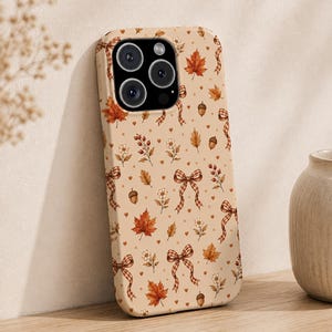 Könnte beinhalten: Eine beige Smartphone-Hülle mit einem wiederkehrenden Muster aus orangefarbenen Ahornblättern, braunen Eicheln, kleinen weißen Blumen, roten Beeren und braunen Vichy-Schleifen. Die Hülle hat einen schwarzen Kameraausschnitt mit drei Linsen.