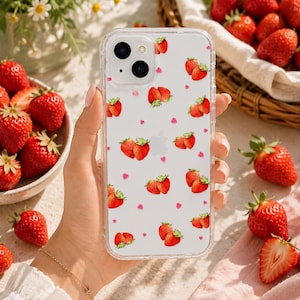 Könnte beinhalten: Transparente Handyhülle mit Erdbeermuster. Die Hülle hat ein sich wiederholendes Muster aus roten Erdbeeren und rosa Herzen auf weißem Hintergrund. Frische Erdbeeren sind um die Hülle herum verstreut.