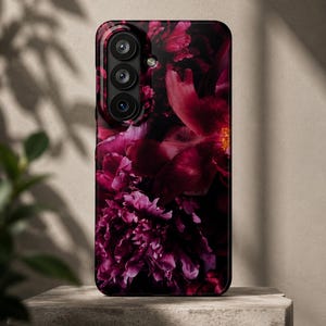 Op de afbeelding: Een rechthoekig smartphonehoesje met een donkere, sfeervolle bloemenprint van diep magenta en bordeauxrode pioenrozen. Het hoesje heeft een verticale camera-uitsparing aan de linkerbovenkant met drie ronde lenzen en een kleine flitser.