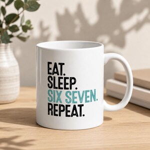 Könnte beinhalten: Weiße Keramik-Tasse mit dem Text "EAT. SLEEP. SIX SEVEN. REPEAT." in schwarzer und teal-farbener Schrift. Die Tasse hat einen gebogenen Henkel und steht auf einer Holzoberfläche. Im Hintergrund ist eine Vase mit Grünpflanzen zu sehen.
