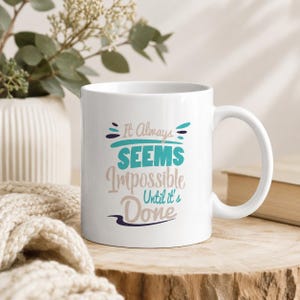 Könnte beinhalten: Ein weißer Keramikbecher mit dem aufgedruckten Text It Always SEEMS Impossible Until it's Done in einer Kombination aus türkisfarbener, beiger und dunkelblauer Schreib- und Blockschrift, mit einem geschwungenen Henkel und glattem Körper.