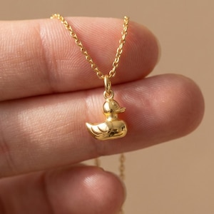 Può includere: Una collana dorata con un ciondolo a forma di anatra. Il ciondolo a forma di anatra è piccolo e dettagliato, sospeso a una sottile catena d'oro. L'immagine mostra la collana su uno sfondo neutro.