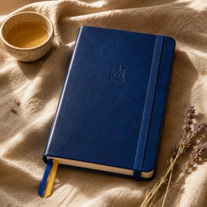 May include: A dark blue notebook with a textured cover featuring an embossed Buddha figure in the center. The notebook includes a vertical blue elastic closure band and two ribbon bookmarks in blue and yellow, extending from the bottom edge.