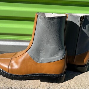 May include: A pair of ankle boots with a two-tone design. The lower portion is tan, while the upper section is gray. The boots have a black sole and a zipper on the side. The boots are made of leather.
