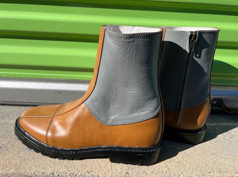 May include: A pair of ankle boots with a two-tone design. The lower portion is tan, while the upper section is gray. The boots have a black sole and a zipper on the side. The boots are made of leather.