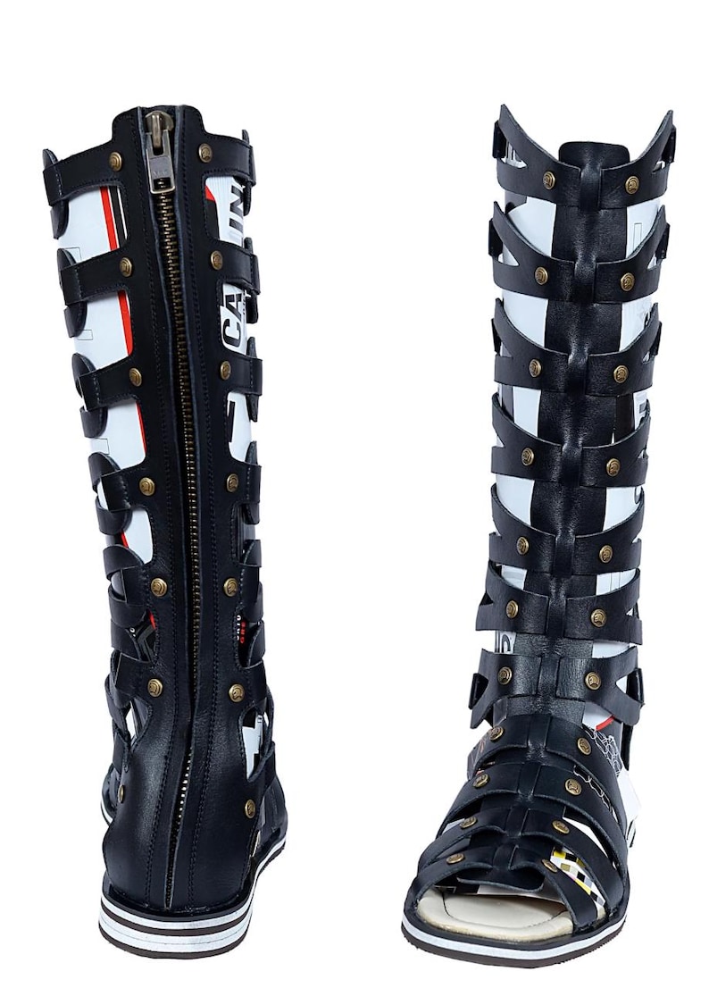 May include: Black and white gladiator sandals with a zipper and strap design. The sandals have a flat sole with white and black stripes. The sandals have the word "CA" printed on the side.