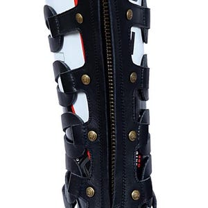 May include: Black and white knee-high gladiator boot with a full-length zipper and gold-colored rivets. The boot features a series of straps and cutouts, with the word "CA" visible on the side.