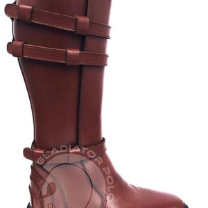 May include: A knee-high brown leather boot featuring two horizontal buckled straps at the upper shaft and one buckled strap at the ankle. The boot has a thick black lug sole and a watermark logo that reads GLADIATOR POLO across the lower side.