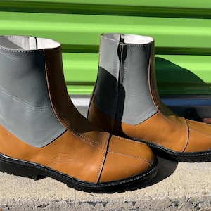 May include: A pair of ankle-high boots with a two-tone design. The boots feature a tan leather lower section and a gray leather upper section. They have black soles and a zipper closure.