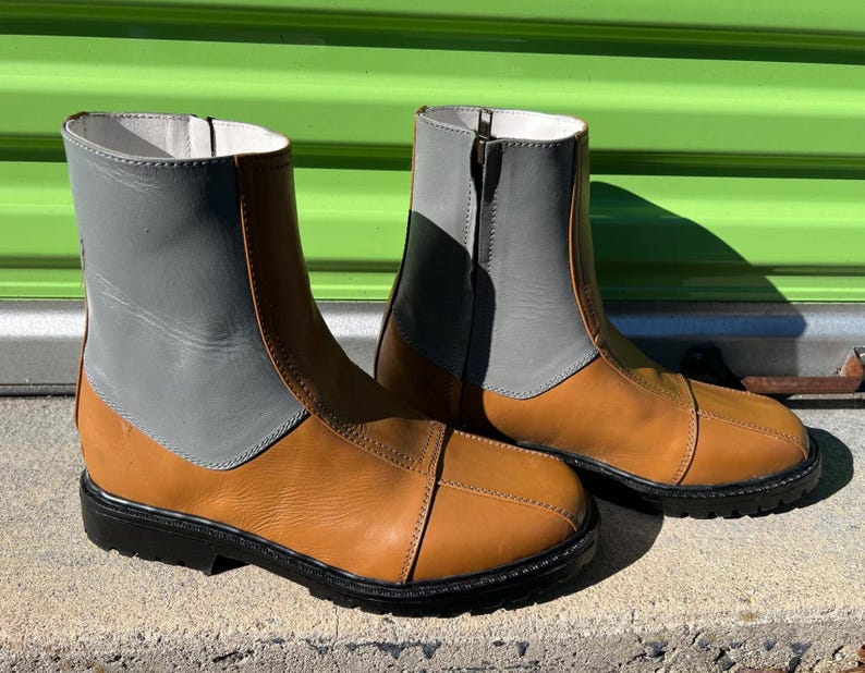 May include: A pair of ankle-high boots with a two-tone design. The boots feature a tan leather lower section and a gray leather upper section. They have black soles and a zipper closure.
