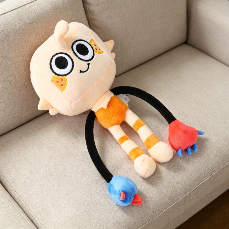 May include: A plush toy with a light peach-colored head featuring large black eyes, a small smile, and two slices of pizza. The toy has orange shorts, striped legs, and black arms ending in red and blue appendages. It is lying on a beige couch.