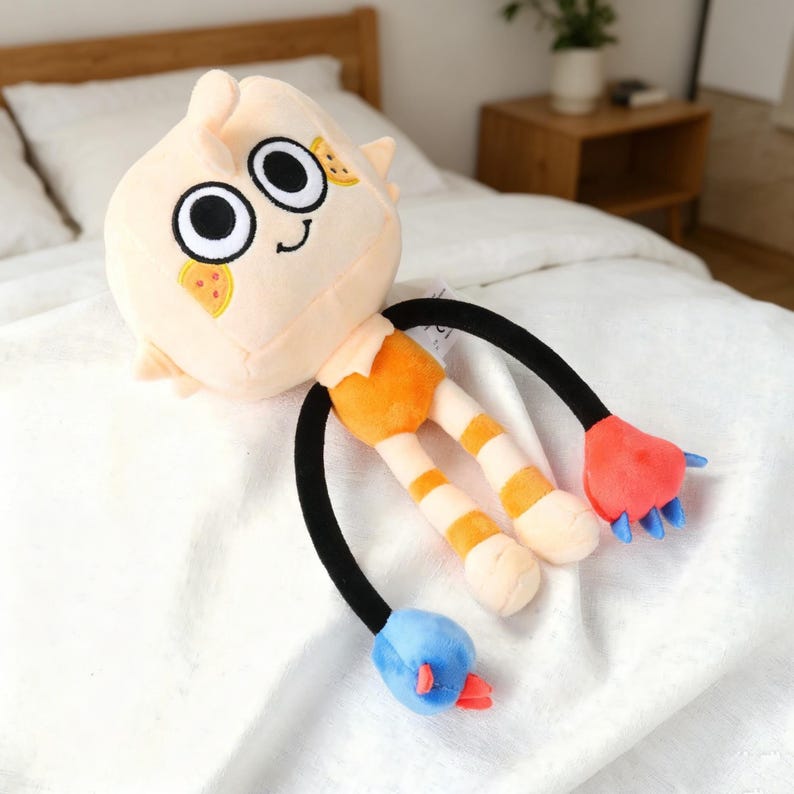 May include: A plush toy with a light peach-colored head and a smiling face with large black eyes. The toy has orange and white striped legs, black arms, and red and blue hand and foot details. The toy is lying on a white surface.