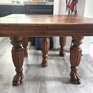 May include: A rectangular, antique wooden table with a dark brown finish. The table features ornate, carved legs with a claw-foot design. The table's edges are detailed with decorative molding, showcasing a classic, vintage style.