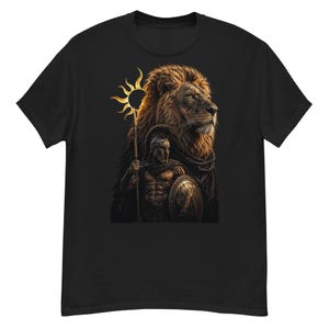 Può includere: Una t-shirt nera a maniche corte con una stampa grafica di un guerriero che indossa un elmo e impugna una lancia e uno scudo, davanti a una grande testa di leone e un simbolo solare dorato. Il disegno è centrato sul petto della maglietta.