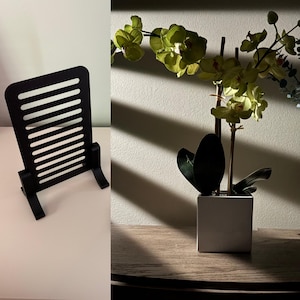 Könnte beinhalten: Ein schwarzer Präsentationsständer mit einer Reihe horizontaler Lamellen und einem stabilen Fuß, neben einer künstlichen Orchidee im Topf mit hellgrünen Blüten und dunkelgrünen Blättern in einem quadratischen weißen Gefäß, beleuchtet durch Streifenschatten.