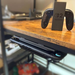 Puede incluir: Un soporte negro para consola y mandos de Nintendo Switch. El soporte sujeta la consola y dos mandos. El logotipo de Nintendo Switch es visible en el soporte. El soporte está sobre un escritorio de madera.