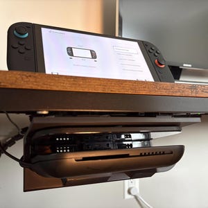 May include: A black video game console with a screen displaying a controller pairing menu, mounted on a dark brown wooden shelf. Below the shelf, a black plastic docking station is secured, holding a secondary console unit in a horizontal position.