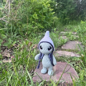 May include: A crocheted doll with a light blue body, wearing a purple hat and purple hair-like strands. The doll has large black eyes with pink highlights. It is standing on a brick in a grassy outdoor setting.