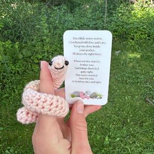 May include: A crocheted pink worry worm with a smiling face, held near a card with text. The card reads, "I'm a little worry worm... So hold me nice and tight."