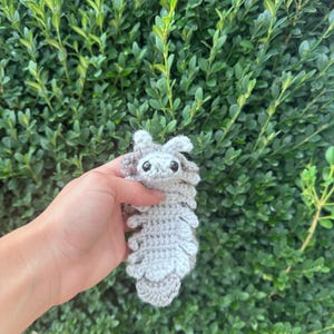 May include: A light grey crocheted isopod plush toy featuring a segmented body, multiple small legs along the sides, two antennae on the head, and two small black plastic safety eyes. The soft yarn creature has a textured surface.