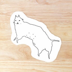 以下が含まれることがあります： 白地に黒い線画で描かれた、横たわる猫のステッカー。猫の輪郭はシンプルで、目やひげなどの小さなディテールがあります。ステッカーは明るい木目調の表面にあります。