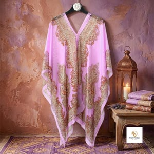 Pode incluir: Um caftan rosa claro com um padrão paisley detalhado em dourado e castanho, decote em V e uma silhueta solta e fluida. A peça é exibida num cabide preto com uma flor branca, destacando os intrincados desenhos ornamentais do tecido.