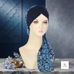 May include: A dark navy blue turban head wrap featuring a pleated front design and long, flowing side panels decorated with a vibrant blue and black floral pattern. The head covering is displayed on a mannequin head with the brand name PASHMIQUE.
