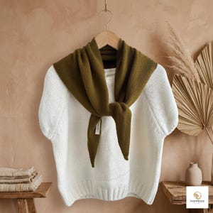 Puede incluir: Un suéter de punto blanco de manga corta con dobladillo y cuello acanalados, combinado con una bufanda de punto triangular de color verde oliva anudada al frente. El suéter presenta una etiqueta de marca blanca visible en el cuello interior y una pequeña etiqueta.