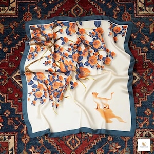 May include: Square silk scarf with a cream background, featuring a floral design in orange and blue. A cat is depicted in the lower right corner. The scarf has a teal border.