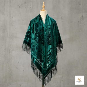 Pode incluir: Um xale de veludo verde esmeralda profundo com um padrão floral de rosas tom sobre tom em todo o tecido. A peça triangular é finalizada com franjas pretas longas e finas nas bordas inferiores. O nome da marca Pashmique está impresso no canto.