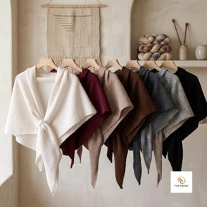 May include: A collection of seven knit shawls hanging on wooden hangers, displayed in a row. The shawls are available in white, burgundy, beige, brown, dark grey, light grey, and black. Each shawl features a front-tie design with pointed ends.