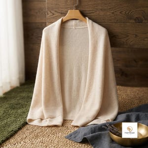 May include: A cream-coloured open-front cardigan with long sleeves and a ribbed hem, hanging on a wooden hanger. The garment features a soft, knit texture and a simple, unfastened design. The brand name PASHMIQUE is visible on a small label.
