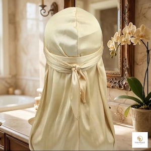 Può includere: Un durag color champagne, in raso, con chiusura annodata. Il durag è drappeggiato, con finiture decorative intorno alla testa e una lunga coda. Lo sfondo mostra un bagno con uno specchio e orchidee.