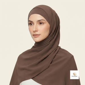 May include: A solid dark brown hijab made of a smooth, opaque fabric that drapes around the head and shoulders. The head covering features a clean, minimalist design with a logo in the bottom corner that reads PASHMIQUE BY HOTA.