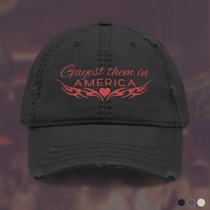 May include: A black distressed baseball cap featuring red embroidered text that reads Gayest them in AMERICA with a small heart and tribal-style flame accents centered below the lettering on the front panel of the hat.