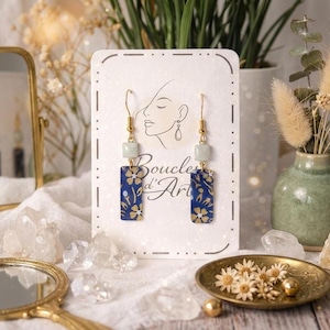 May include: Gold-tone earrings with a rectangular blue pendant featuring a gold floral pattern. Each earring has a small, light green square bead and a gold hook. The earrings are displayed on a white card with the text "Boucles d'Art".