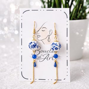 Può includere: Orecchini dorati con perline in ceramica a motivi floreali bianchi e blu. Gli orecchini hanno ciondoli blu a forma di goccia e catenine dorate. La carta dietro gli orecchini ha il testo "Boucles d'Art" e un disegno a tratto del volto di una donna.
