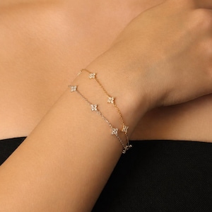 May include: A gold and silver bracelet with a floral design is worn on a wrist. The bracelet features small, flower-shaped charms. The skin tone is light, and the person is wearing a black top.
