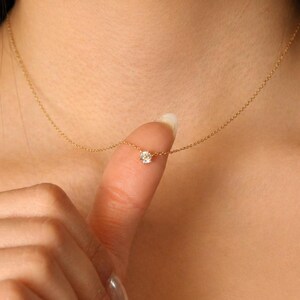 May include: A delicate gold necklace featuring a small, round, clear gemstone pendant. The necklace is shown on a person's skin, with the pendant resting on a fingertip. The chain is fine and the gemstone sparkles.