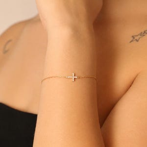 May include: A gold bracelet featuring a cross pendant set with small, sparkling stones. The delicate bracelet is worn on the wrist. The background shows a person's shoulder and arm. The cross pendant is a religious symbol.