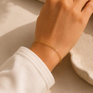 May include: A delicate gold chain bracelet featuring small, elongated rectangular links. The bracelet is worn on a wrist, showcasing a minimalist design with a polished finish that catches the light, creating a subtle and refined appearance on the skin.