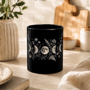 May include: A black ceramic mug featuring a white graphic print of the lunar cycle phases interspersed with detailed botanical illustrations of wildflowers, ferns, and small stars, wrapping around the exterior surface of the cylindrical vessel.