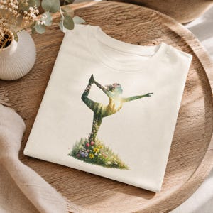 Könnte beinhalten: Ein cremefarbenes T-Shirt mit einem Grafikdruck einer Person in einer Yoga-Pose. Die Silhouette ist mit einer Waldlandschaft, Sonnenlicht und einer Basis aus bunten Wildblumen gefüllt, darunter rosa, gelbe und weiße Blüten auf grünem Grund.