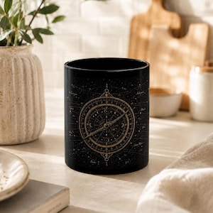 Könnte beinhalten: Schwarze Keramik-Tasse mit detailliertem goldenen Kompass-Design vor sternenklarem Nachthimmel. Die zylindrische Tasse steht in einer neutralen Küchenumgebung mit einer Vase mit Grünpflanzen.