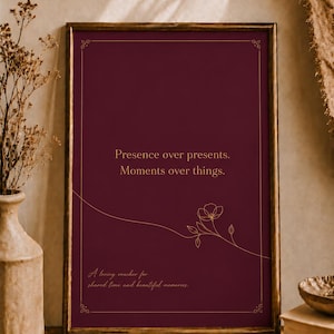 Könnte beinhalten: Gerahmter burgunderfarbener Druck mit goldenem Text und floralem Design. Der Text lautet "Presence over presents. Moments over things." und "A loving voucher for shared time and beautiful memories."