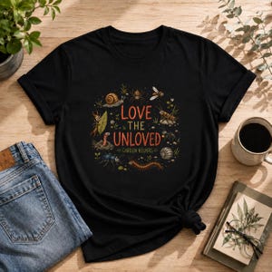 Könnte beinhalten: Ein schwarzes T-Shirt mit kurzen Ärmeln und einem zentralen Aufdruck verschiedener Garteninsekten und Kleintiere, die den Text "LOVE THE UNLOVED GARDEN HELPERS" in orangefarbenen und weißen Buchstaben umgeben, darunter eine Schnecke und eine Spinne.