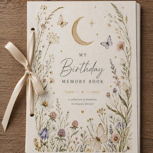 Könnte beinhalten: Ein cremefarbenes Erinnerungsbuch mit dem Titel MY Birthday MEMORY BOOK, verziert mit einer Mondsichel, Sternen und Schmetterlingen. Das Cover zeigt Aquarell-Wildblumen und Gräser, gebunden mit einer beigen Satinschleife an der linken Seite.