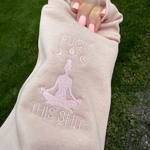 May include: A light pink sweatshirt featuring embroidered text that reads FUCK THIS SHIT above a silhouette of a person sitting in a lotus meditation pose, with small moon phase symbols embroidered between the text and the figure.